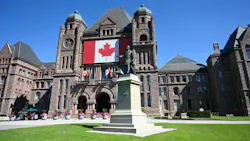 The Ontario Legislative Building at Queen's Park in Toronto, its sandstone facade draped with a large Canadian flag, with a statue of former premier Whitney in the foreground and flowering planters lining the entrance.