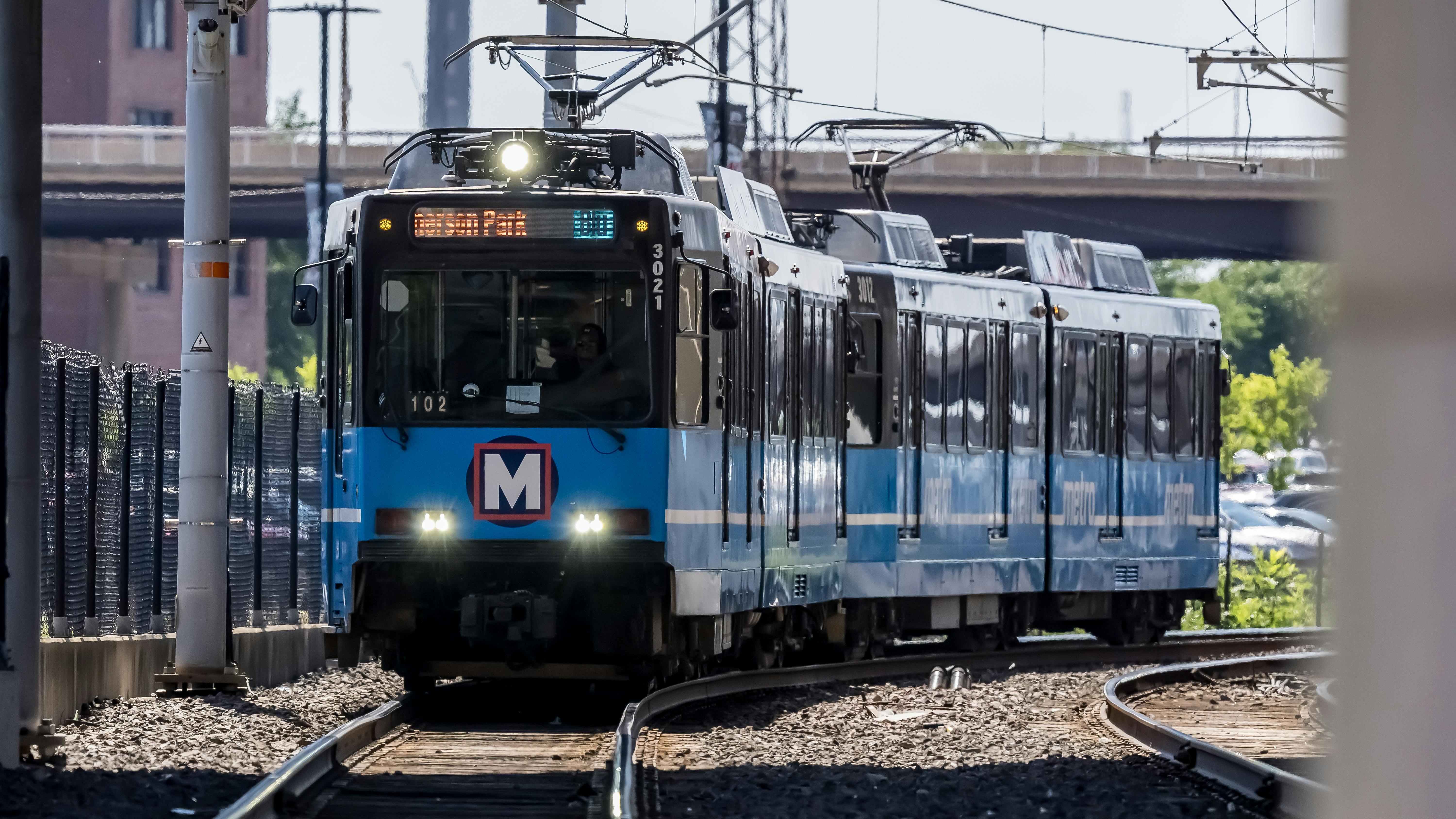A Metro light rail train, numbered 3021 and signed for Merson Park, approaches on curved tracks beneath a bridge, with its 'M' logo visible on the front and a chain-link fence lining the tracks.