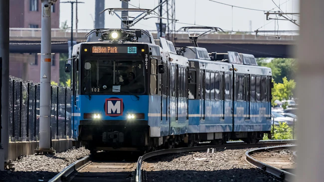 A Metro light rail train, numbered 3021 and signed for Merson Park, approaches on curved tracks beneath a bridge, with its 'M' logo visible on the front and a chain-link fence lining the tracks.