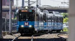 A Metro light rail train, numbered 3021 and signed for Merson Park, approaches on curved tracks beneath a bridge, with its 'M' logo visible on the front and a chain-link fence lining the tracks.