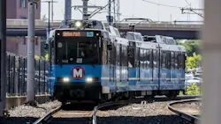 A Metro light rail train, numbered 3021 and signed for Merson Park, approaches on curved tracks beneath a bridge, with its 'M' logo visible on the front and a chain-link fence lining the tracks.