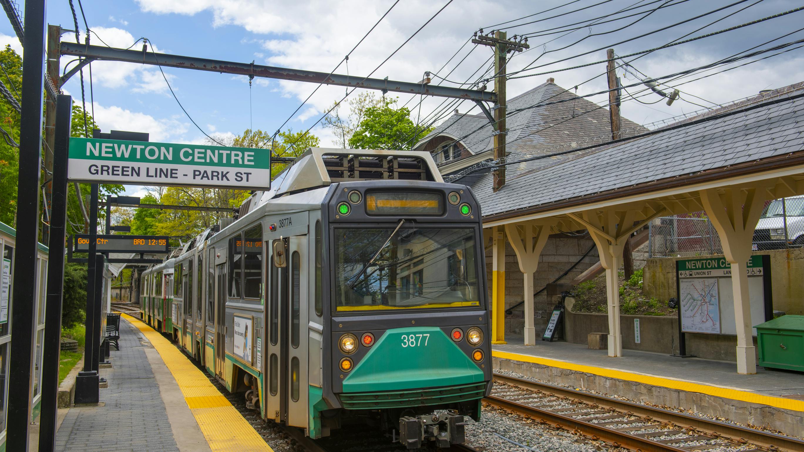 An MBTA Green Line trolley, car 3877, arrives at the Newton Centre station on a sunny day, with the historic station building visible alongside the platform.