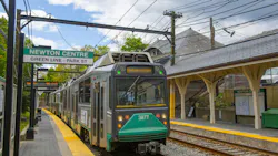 An MBTA Green Line trolley, car 3877, arrives at the Newton Centre station on a sunny day, with the historic station building visible alongside the platform.