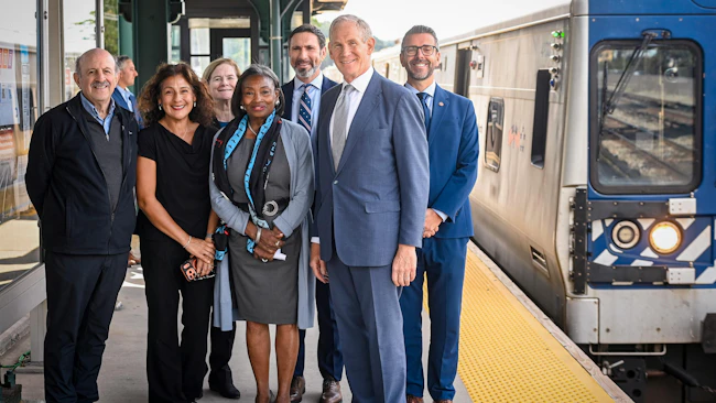A group of seven officials pose on a Metro-North Railroad platform beside a stopped train, smiling for a photo under the station canopy.