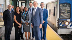 A group of seven officials pose on a Metro-North Railroad platform beside a stopped train, smiling for a photo under the station canopy.