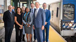 A group of seven officials pose on a Metro-North Railroad platform beside a stopped train, smiling for a photo under the station canopy.