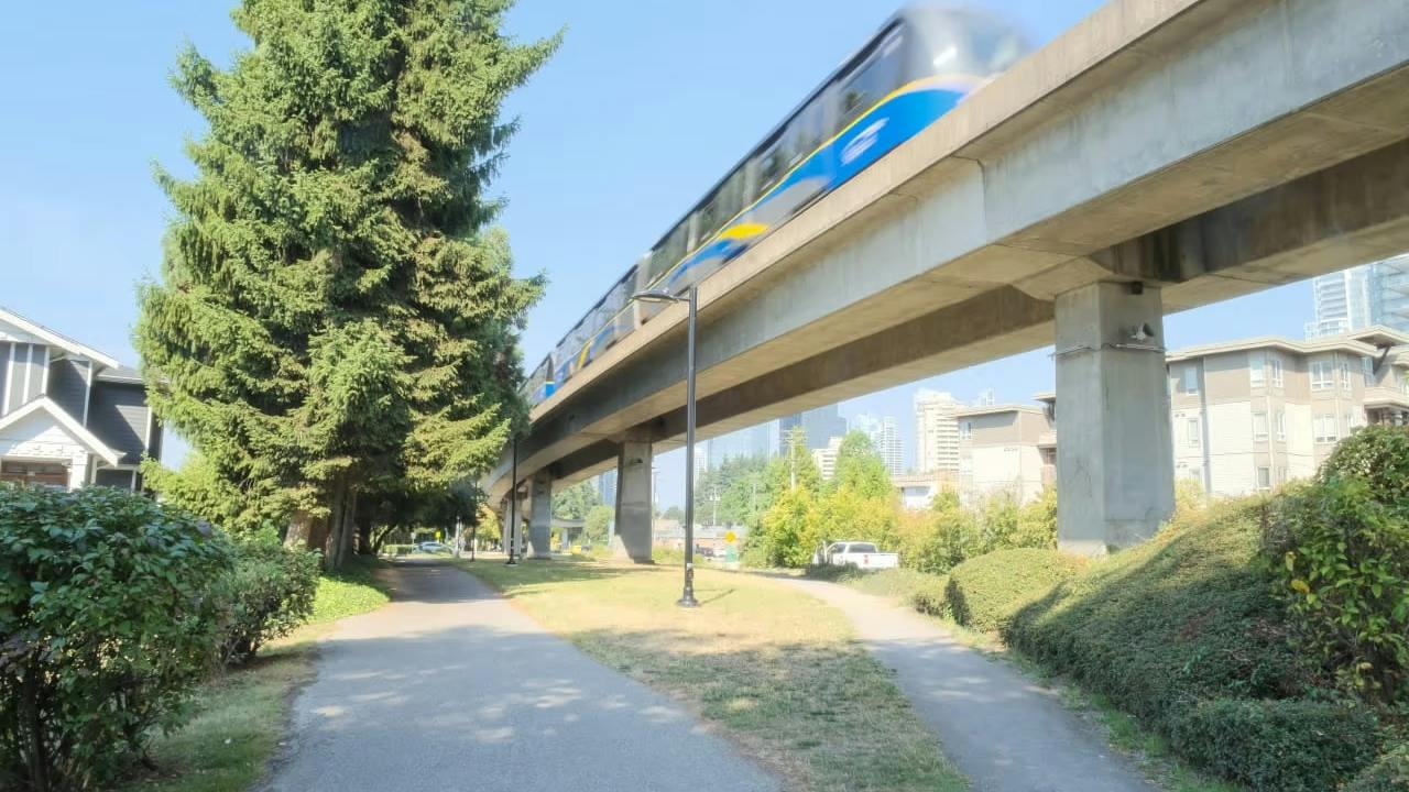 A SkyTrain train travels along an elevated guideway above a tree-lined pathway, with residential buildings and a distant city skyline visible in the background.