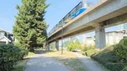A SkyTrain train travels along an elevated guideway above a tree-lined pathway, with residential buildings and a distant city skyline visible in the background.