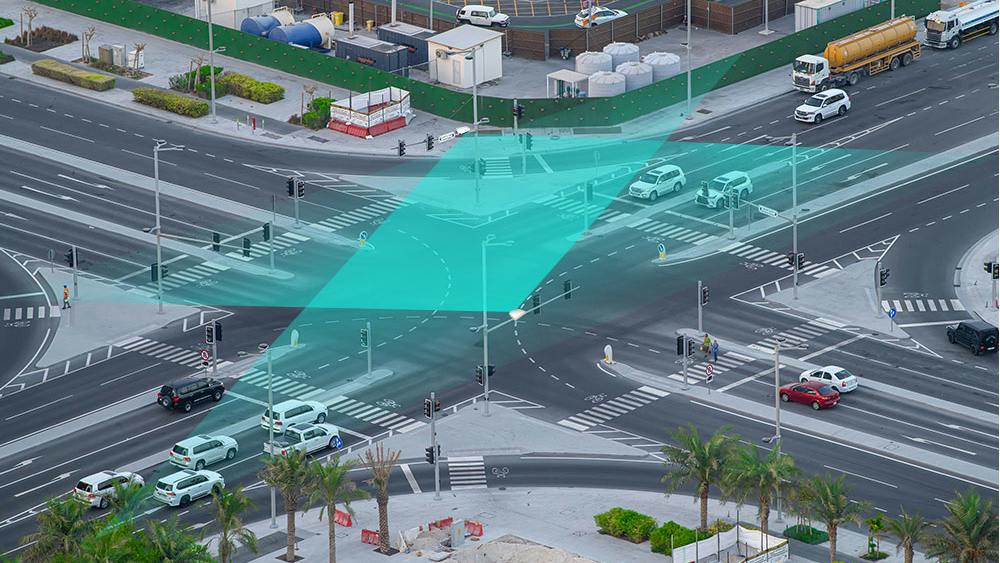 An aerial view of a busy intersection shows a teal-colored graphic overlay illustrating vehicle detection and signal timing technology used to coordinate traffic flow.