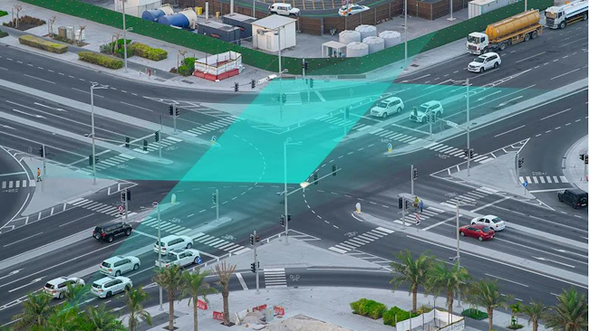 An aerial view of a busy intersection shows a teal-colored graphic overlay illustrating vehicle detection and signal timing technology used to coordinate traffic flow.