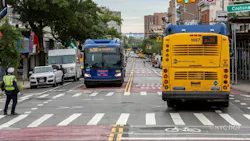 A traffic enforcement agent watches from a Bronx crosswalk as an MTA New York City Bus and a Bx36 bus pass each other in dedicated bus lanes marked with red pavement and bike symbols, with a 'Crotona' street sign and pedestrian signals overhead.