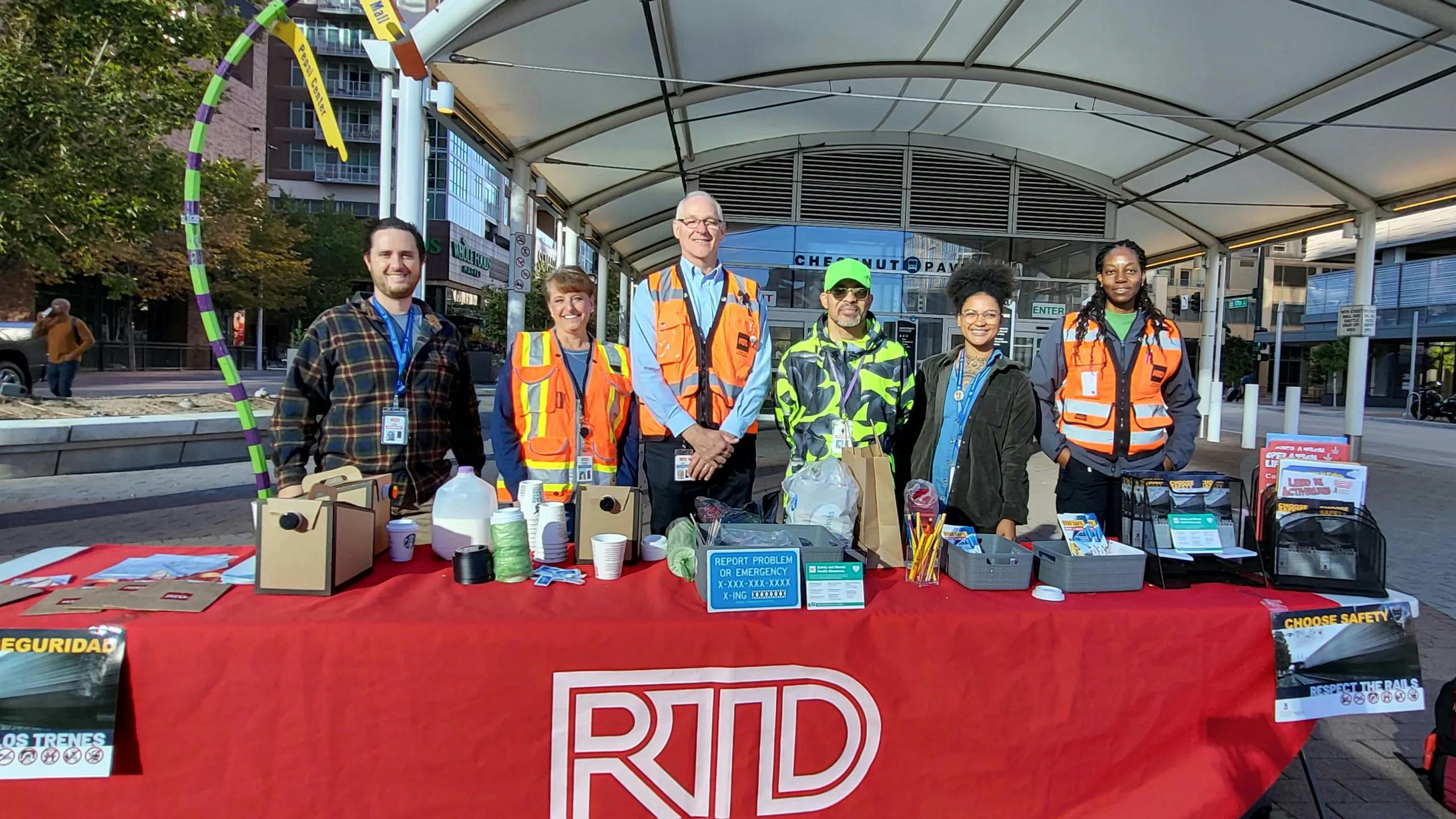 Six Denver RTD employees stand behind a red table branded with the agency logo, displaying rail safety materials in English and Spanish at an outdoor transit plaza.