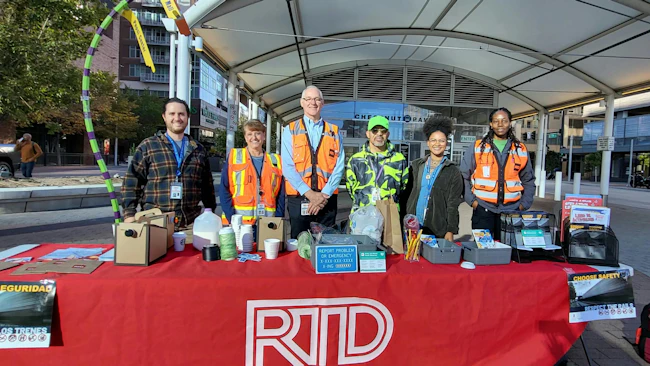 Six Denver RTD employees stand behind a red table branded with the agency logo, displaying rail safety materials in English and Spanish at an outdoor transit plaza.