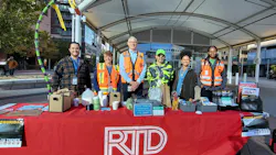 Six Denver RTD employees stand behind a red table branded with the agency logo, displaying rail safety materials in English and Spanish at an outdoor transit plaza.
