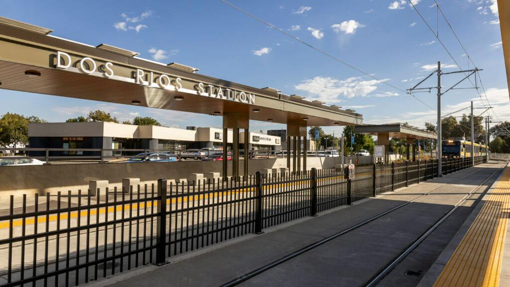 The newly opened Dos Rios light-rail station, with an overhead canopy displaying the station name, a light-rail train approaching on the tracks, and a black metal fence lining the platform.