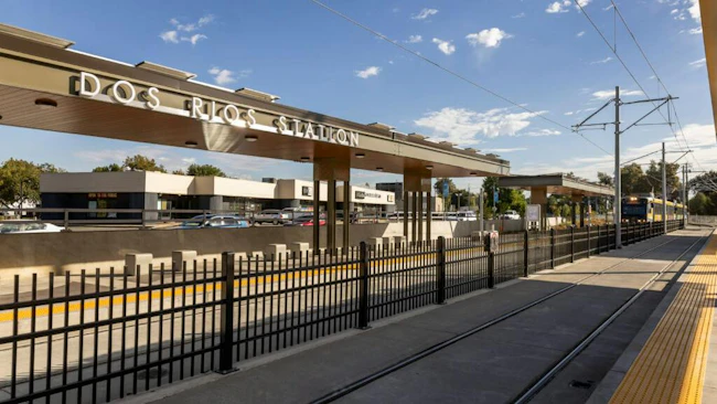 The newly opened Dos Rios light-rail station, with an overhead canopy displaying the station name, a light-rail train approaching on the tracks, and a black metal fence lining the platform.