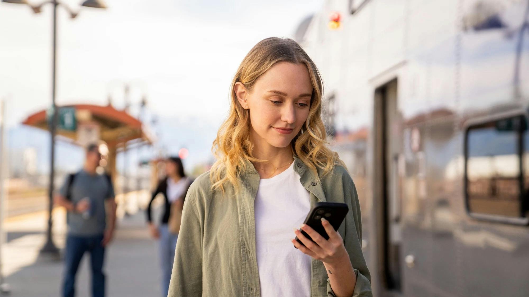 A woman waiting at a train platform checks her phone, illustrating Metrolink's rollout of a new version of the See Say ELERTS safety-reporting app, with fellow passengers and a train visible in the background.