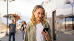 A woman waiting at a train platform checks her phone, illustrating Metrolink's rollout of a new version of the See Say ELERTS safety-reporting app, with fellow passengers and a train visible in the background.
