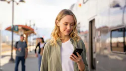 A woman waiting at a train platform checks her phone, illustrating Metrolink's rollout of a new version of the See Say ELERTS safety-reporting app, with fellow passengers and a train visible in the background.