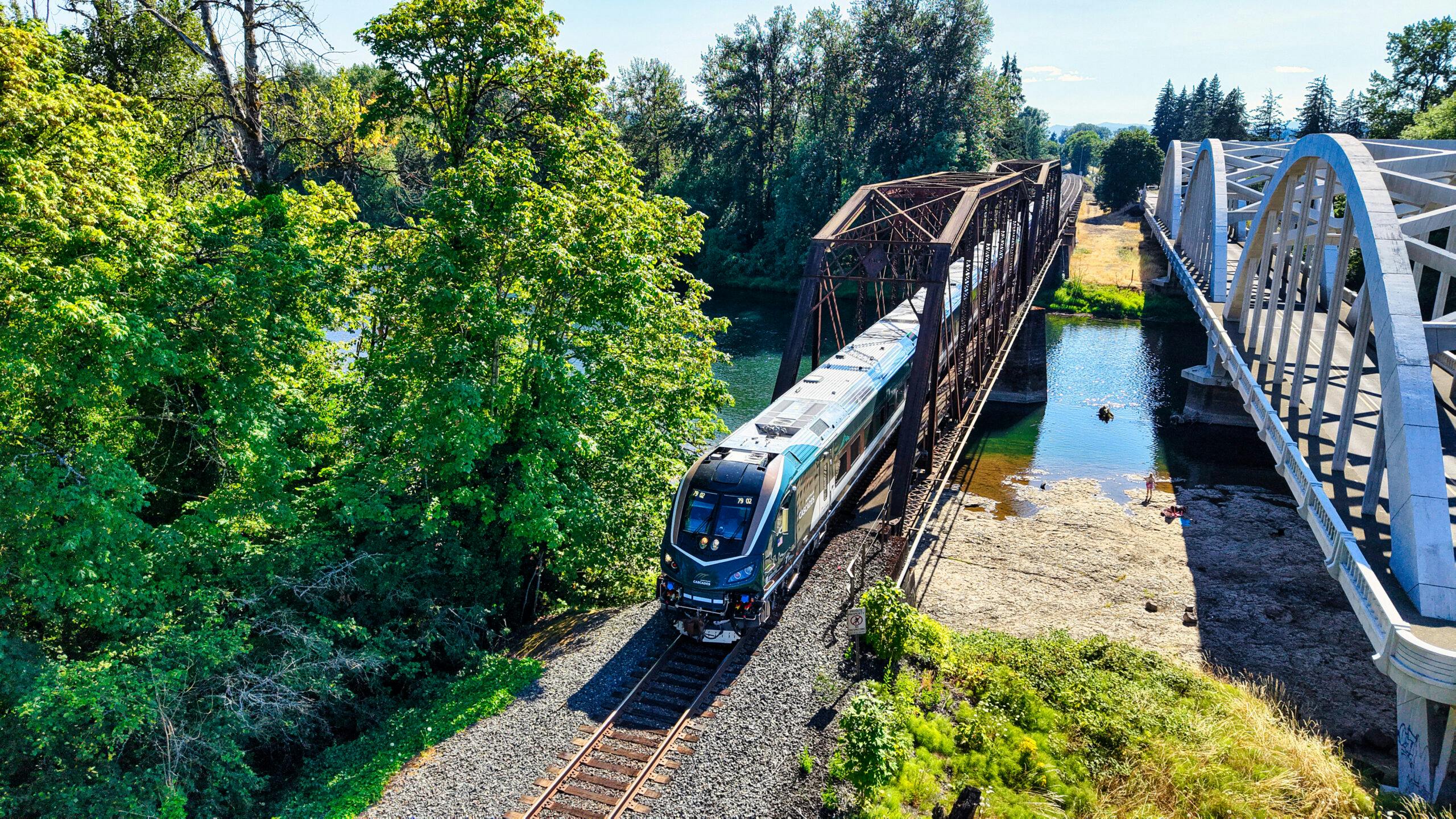 An Amtrak Cascades train, locomotive 79 02, crosses a steel truss bridge over a river alongside a white arched pedestrian/vehicle bridge in a wooded setting.