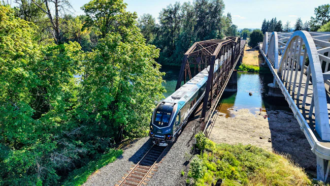 An Amtrak Cascades train, locomotive 79 02, crosses a steel truss bridge over a river alongside a white arched pedestrian/vehicle bridge in a wooded setting.