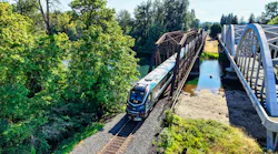 An Amtrak Cascades train, locomotive 79 02, crosses a steel truss bridge over a river alongside a white arched pedestrian/vehicle bridge in a wooded setting.