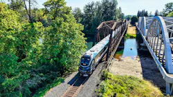 An Amtrak Cascades train, locomotive 79 02, crosses a steel truss bridge over a river alongside a white arched pedestrian/vehicle bridge in a wooded setting.