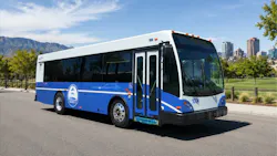 A blue and white Minot City Transit bus, numbered 1004, parked in a lot with a city skyline and mountains visible in the background.
