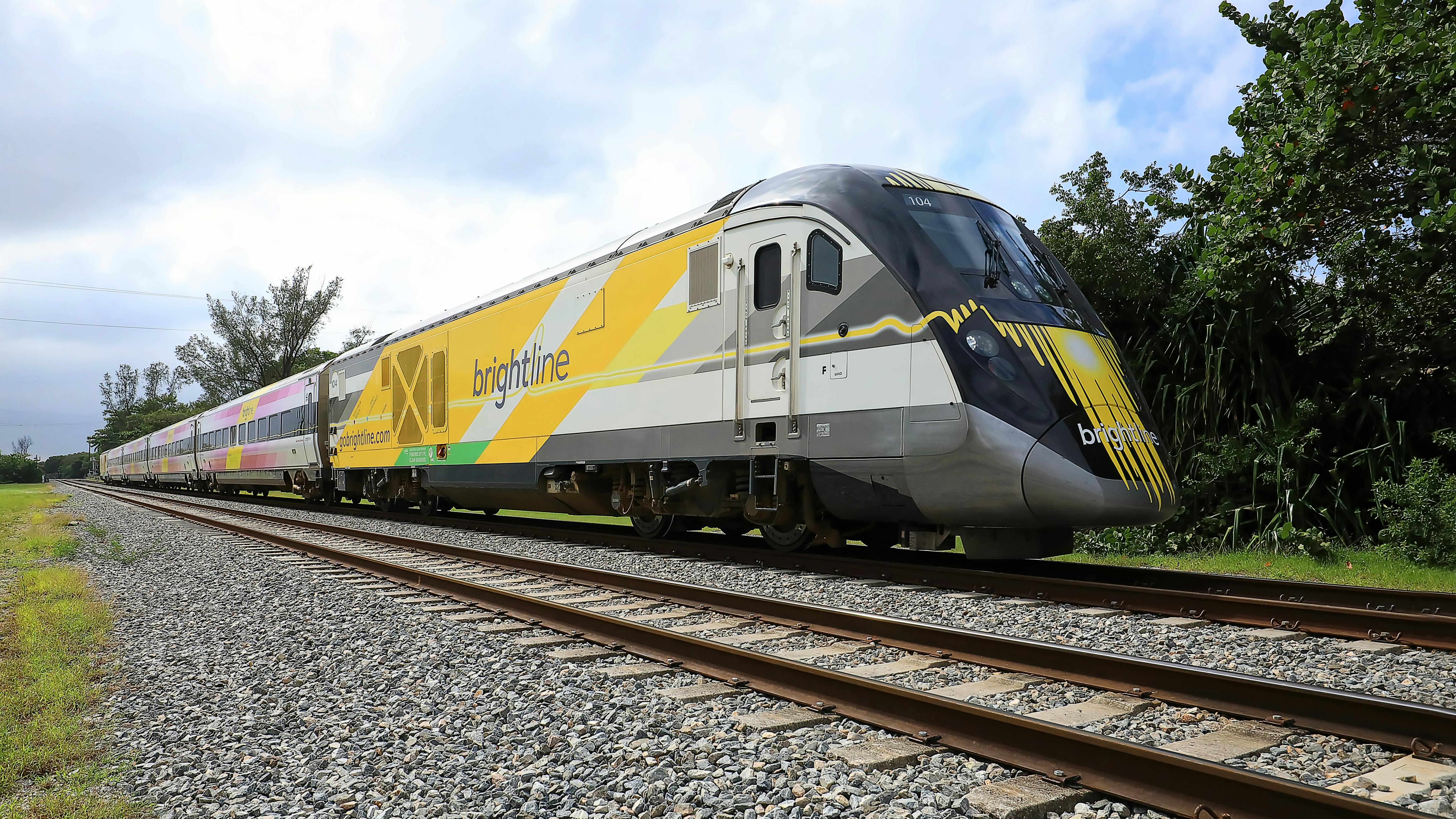 A Brightline high-speed train, numbered 104, travels along tracks lined with gravel and trees, displaying the company's yellow, gray and black livery.