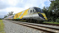 A Brightline high-speed train, numbered 104, travels along tracks lined with gravel and trees, displaying the company's yellow, gray and black livery.