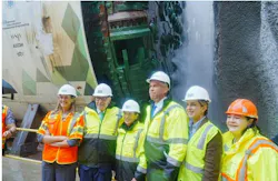 Officials in hard hats and safety vests pose near a tunnel boring machine marked with Gateway Program Trans-Hudson Partnership Engineering branding as tunnel boring begins.