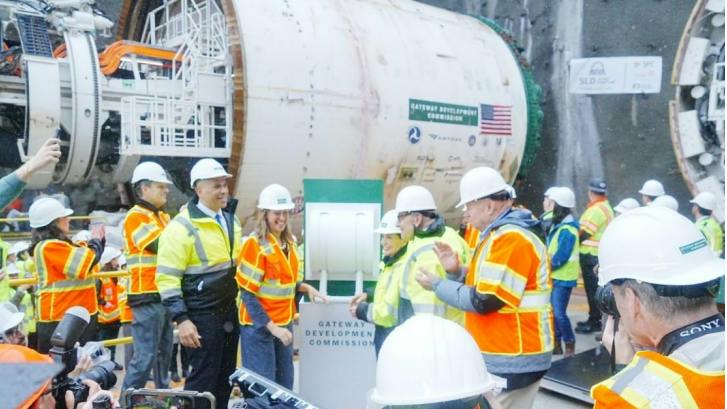 A crowd of officials and media gathers around a large tunnel boring machine marked with Gateway Development Commission branding, with someone about to press a ceremonial button to launch boring.