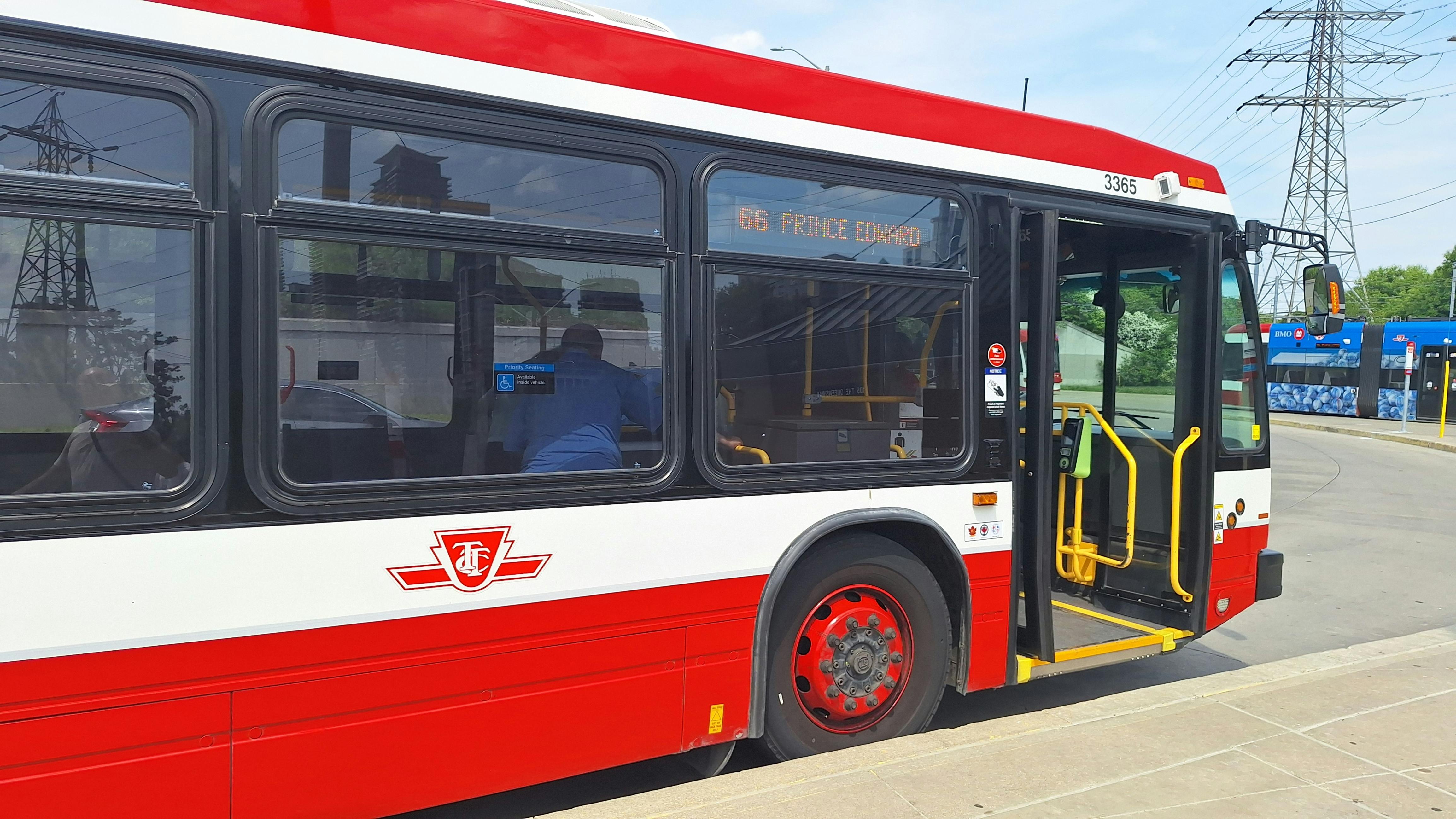 A TTC bus, numbered 3365 and signed '66 Prince Edward,' stands with its doors open at a curb, showing the agency's logo and priority seating signage, with a streetcar and power transmission towers visible in the background.