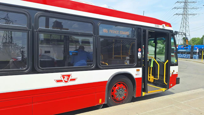 A TTC bus, numbered 3365 and signed '66 Prince Edward,' stands with its doors open at a curb, showing the agency's logo and priority seating signage, with a streetcar and power transmission towers visible in the background.