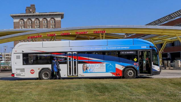 A GCRTA 'Cleaner Air Express' bus, unit 3358, is parked at a transit center in Cleveland with a passenger boarding, under a canopy structure and blue sky.