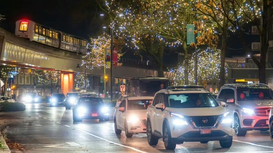 Cars with headlights on drive along a rain-slicked city street at night, passing beneath an elevated SkyTrain guideway and trees strung with holiday lights.