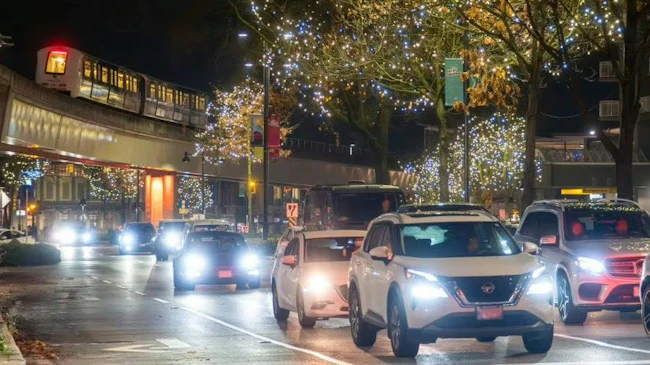 Cars with headlights on drive along a rain-slicked city street at night, passing beneath an elevated SkyTrain guideway and trees strung with holiday lights.