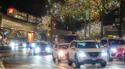 Cars with headlights on drive along a rain-slicked city street at night, passing beneath an elevated SkyTrain guideway and trees strung with holiday lights.