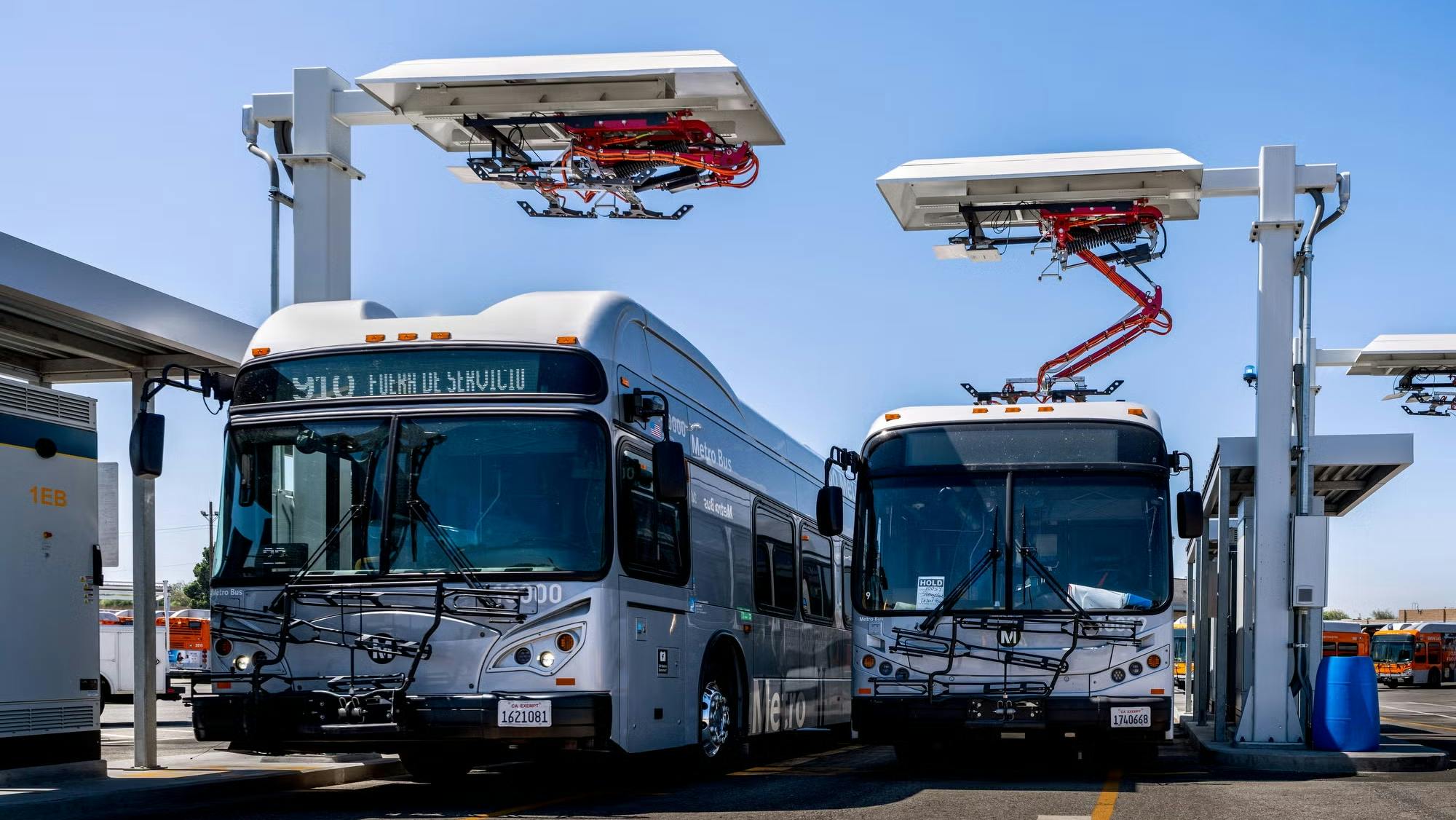 Two L.A. Metro electric buses are parked beneath overhead pantograph charging stations at El Monte Station in California.