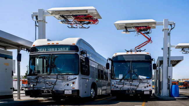Two L.A. Metro electric buses are parked beneath overhead pantograph charging stations at El Monte Station in California.