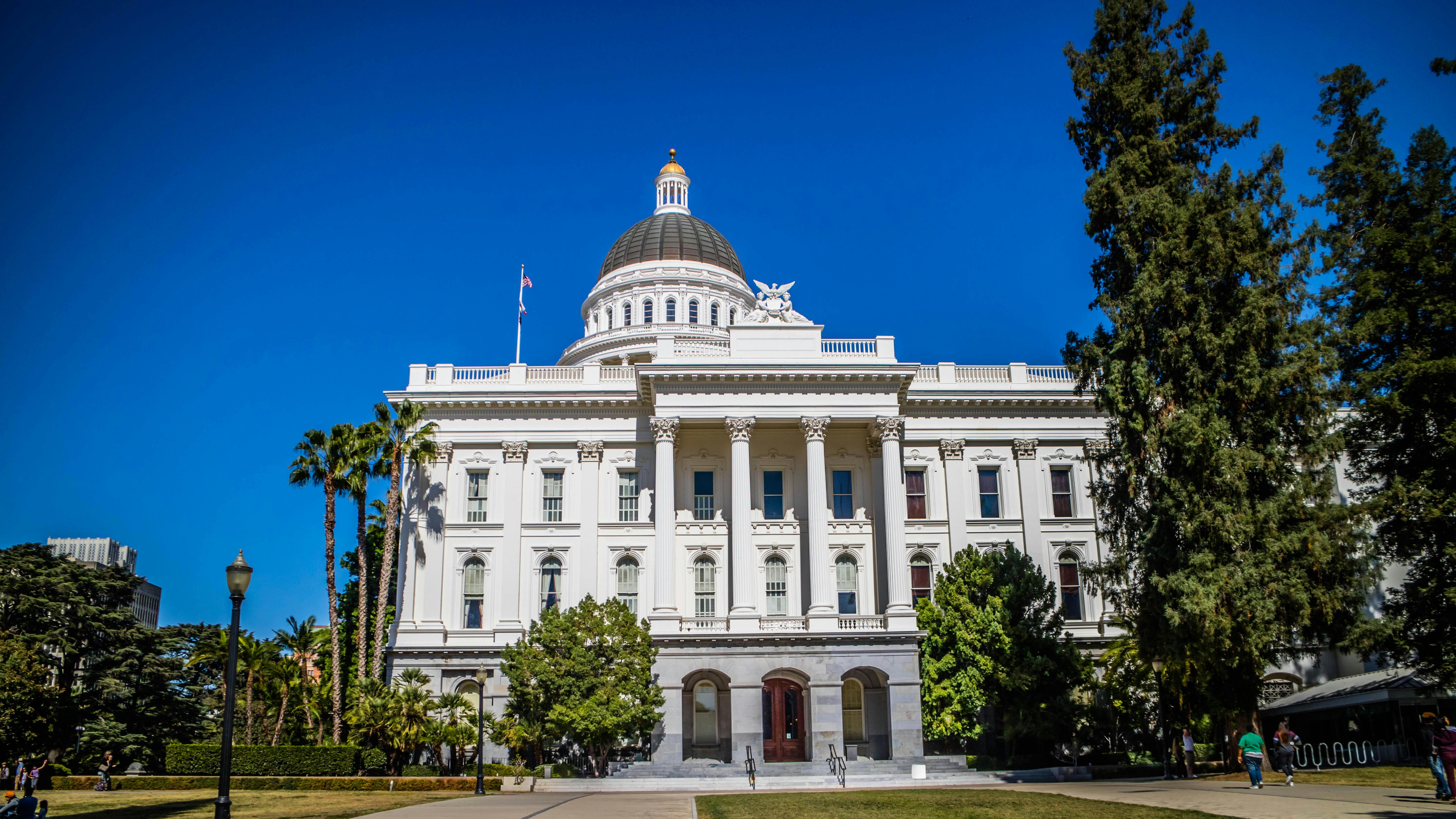 The California State Capitol building in Sacramento, showing its white Corinthian columns, domed rotunda topped with a gold finial, and surrounding palm trees and lawn.