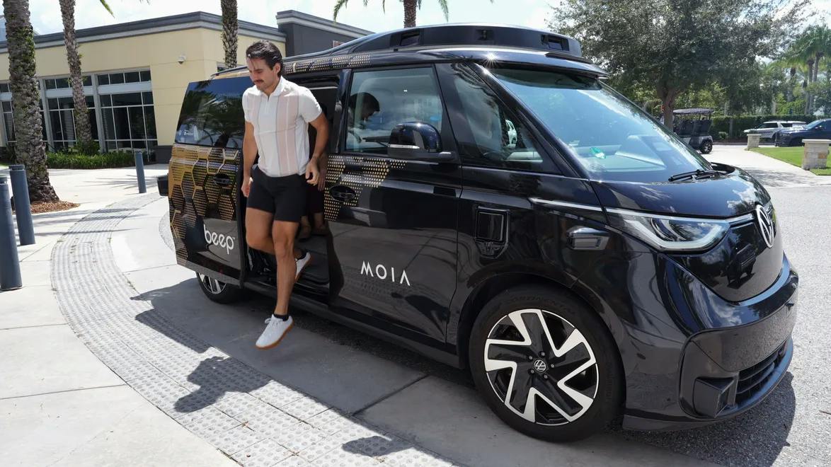 A man exits a black Beep/MOIA autonomous shuttle, based on a Volkswagen ID. Buzz, parked at a curb lined with palm trees and golf carts.