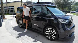 A man exits a black Beep/MOIA autonomous shuttle, based on a Volkswagen ID. Buzz, parked at a curb lined with palm trees and golf carts.