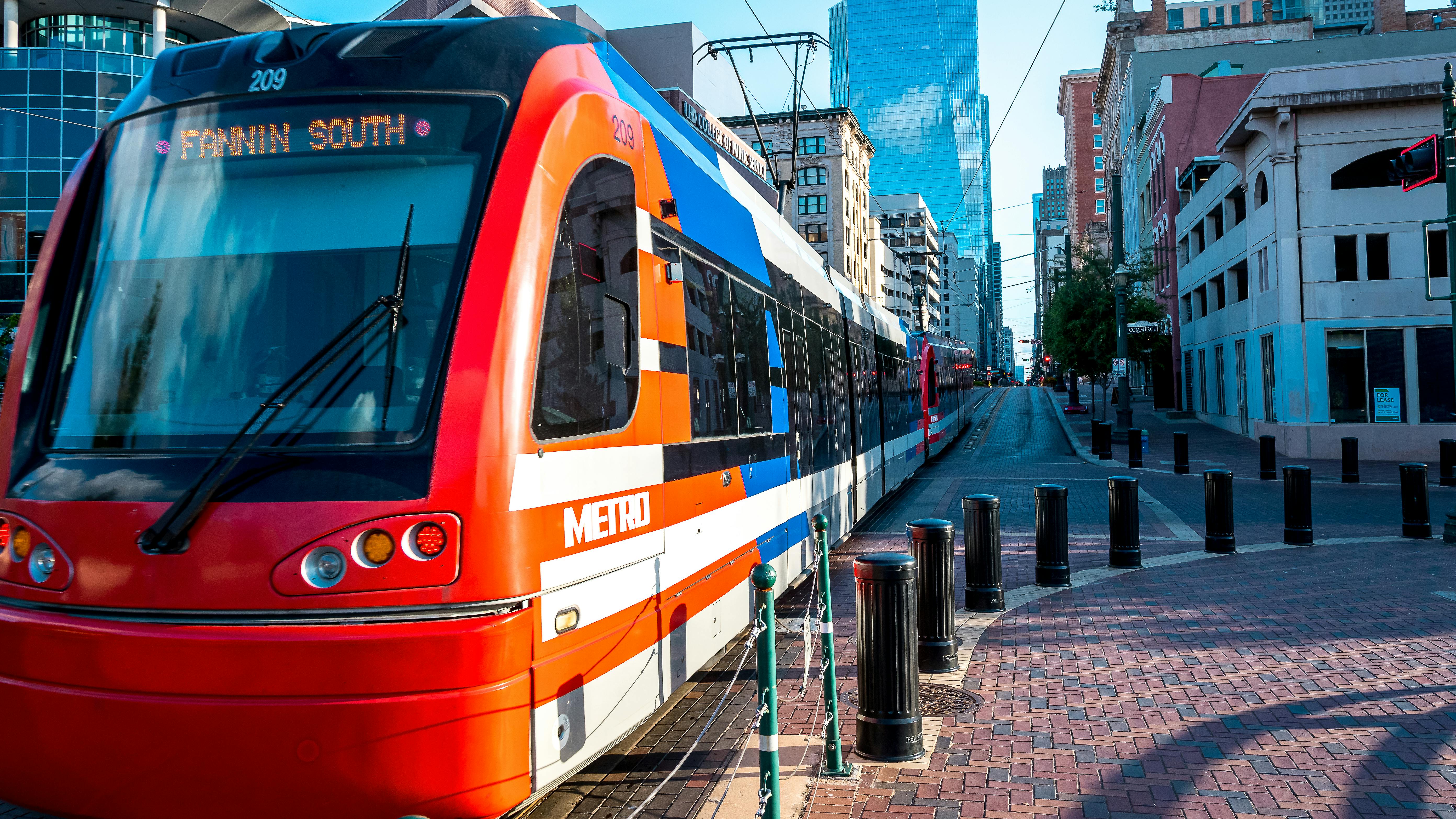 A Houston Metro Rail light rail train, numbered 209 and signed 'Fannin South,' travels down a brick-paved downtown street lined with historic and modern buildings.
