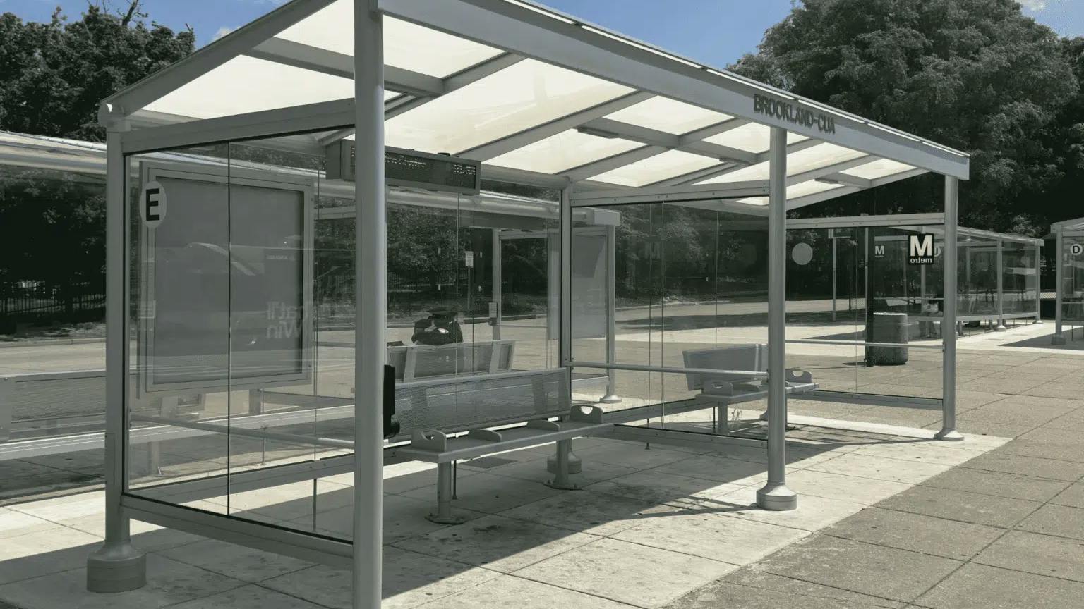 A photo of a new glass-and-steel bus shelter at WMATA's Brookland-CUA station, with digital displays, benches, and the Metro 'M' logo visible, set against a backdrop of trees rendered in color.