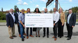 Nine people pose outdoors holding an oversized Amtrak check for $4.1 million made out to West & Central Baltimore Communities for Community Investment Program Grants, with rail catenary structures in the background.