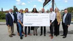 Nine people pose outdoors holding an oversized Amtrak check for $4.1 million made out to West & Central Baltimore Communities for Community Investment Program Grants, with rail catenary structures in the background.