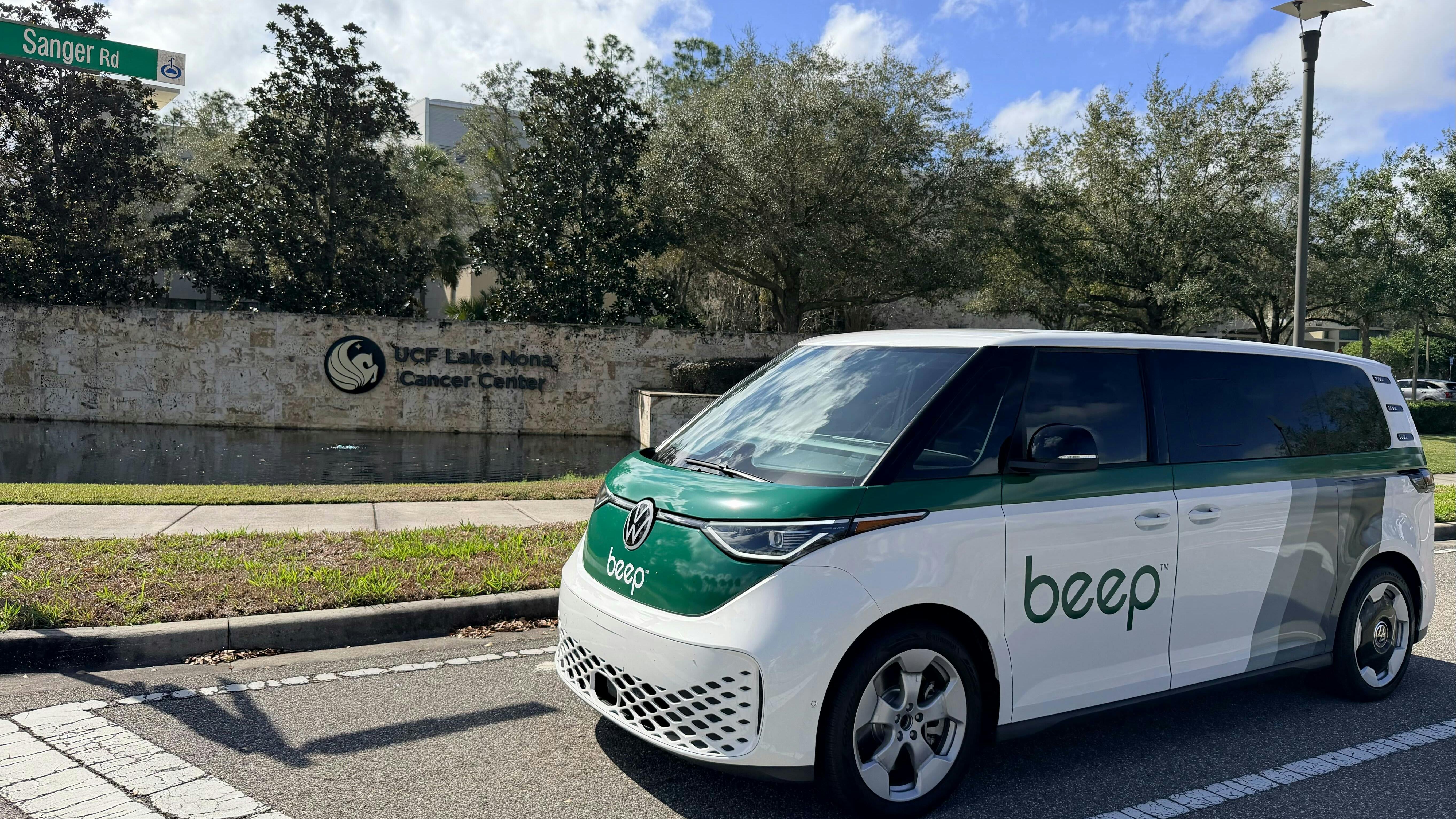 A white and green Beep-branded Volkswagen ID. Buzz van is parked on a street in front of the UCF Lake Nona Cancer Center sign, with a Sanger Road street sign visible above.
