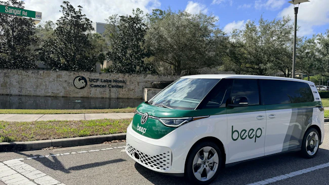 A white and green Beep-branded Volkswagen ID. Buzz van is parked on a street in front of the UCF Lake Nona Cancer Center sign, with a Sanger Road street sign visible above.