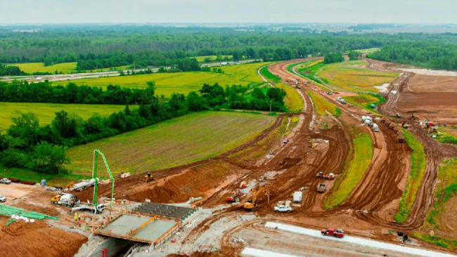 An aerial view of construction on the MetroLink extension to MidAmerica St. Louis Airport shows graded dirt roadbed, a concrete underpass taking shape and heavy equipment surrounded by farmland and trees.
